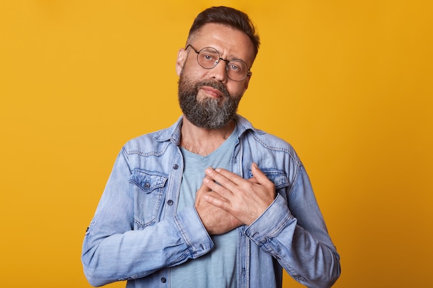 Imagen de un hombre barbudo complacido con lentes ópticos redondeados, mantiene las manos en el pecho, viste una chaqueta de mezclilla de moda