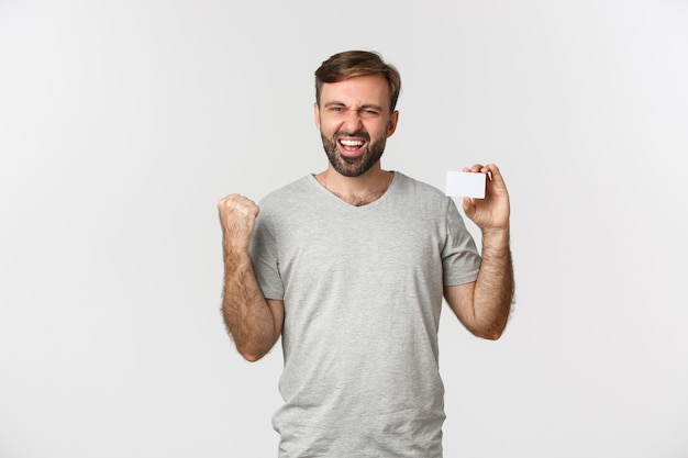 Imagen de hombre barbudo emocionado en camiseta gris, mostrando tarjeta de crédito y haciendo bomba de puño, diciendo que sí