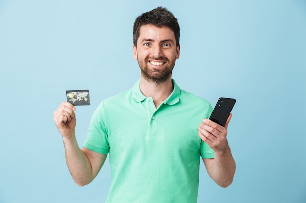 Imagen de hombre barbudo guapo joven emocionado posando aislado sobre pared azul con tarjeta de crédito mediante teléfono móvil.