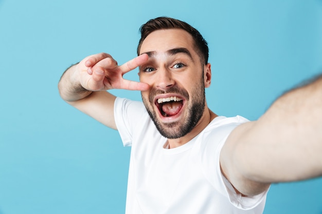 Imagen de hombre barbudo joven alegre posando aislado sobre pared azul tomar un selfie con cámara mostrando gesto de paz.