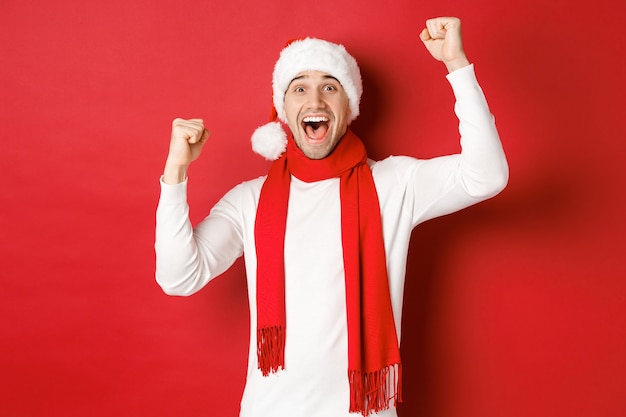 Imagen de hombre caucásico alegre con sombrero de santa y bufanda gritando de alegría y levantando las manos celebrando ...