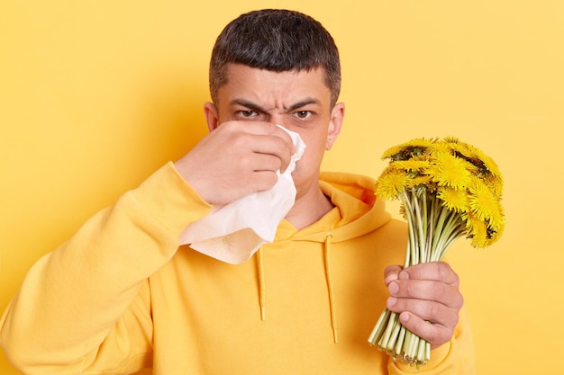 Imagen de un hombre enfermo con capucha de estilo informal posando aislado sobre fondo amarillo sosteniendo un ramo de dientes de león y un pañuelo que sufre alergia