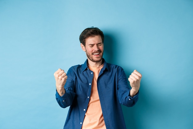 Imagen de hombre feliz celebrando el éxito, gritando sí y agitando los puños cerrados, logrando la meta y triunfando, ganando y bailando, de pie sobre fondo azul.