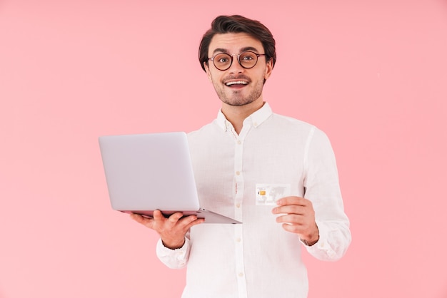 Imagen de hombre feliz guapo con anteojos sosteniendo una computadora portátil y una tarjeta de crédito mientras sonríe