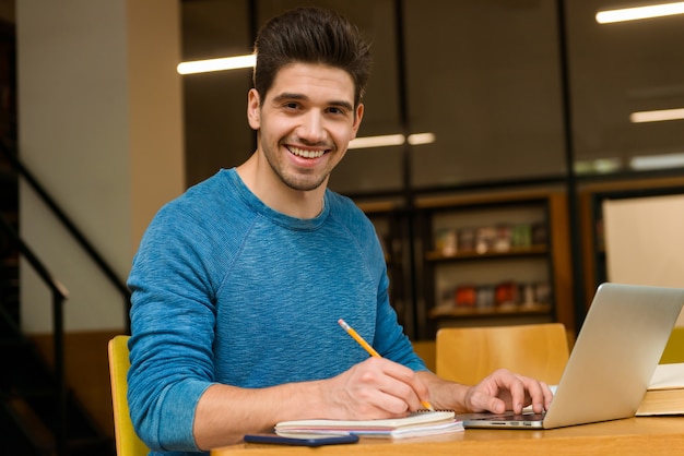 Imagen de un hombre feliz joven estudiante en la biblioteca haciendo la tarea estudiando leer y usar la computadora portátil.