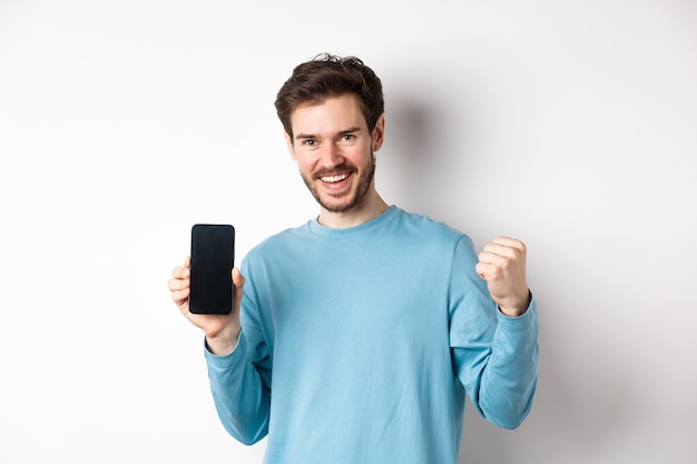 Imagen de hombre feliz mostrando la pantalla vacía del teléfono inteligente y celebrando, sonriendo con regocijo y bomba de puño, lograr el objetivo en línea, de pie sobre fondo blanco.