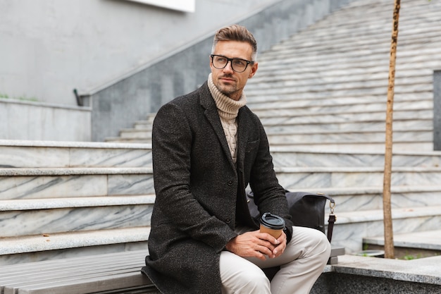 Imagen de hombre guapo de 30 años con anteojos, sentado en las escaleras con café para llevar mientras pasea por las calles de la ciudad