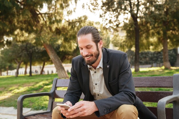 Imagen de hombre guapo con barba de 30 años en traje formal sosteniendo y usando el teléfono móvil, mientras está sentado en un banco en el parque de la ciudad durante el día soleado