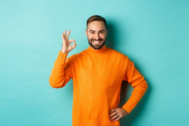 Imagen de hombre guapo confiado que muestra el signo ok, aprobar y estar de acuerdo, como algo bueno, de pie en un suéter naranja.