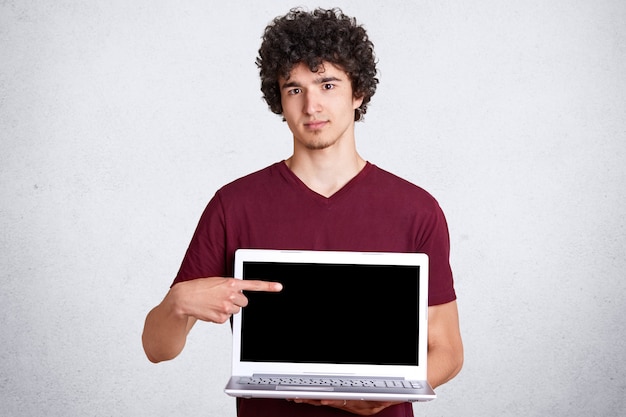 La imagen del hombre hermoso joven en la camiseta marrón casual, se coloca con la computadora portátil muestra con el dedo índice en la pantalla vacía aislada en la pared blanca, espacio libre para su contenido publicitario o promoción.