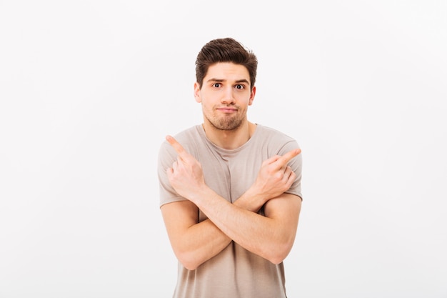 Imagen del hombre indeciso con camiseta beige gesticulando con los dedos a un lado con los brazos cruzados en dos opciones de copyspace, aislado sobre la pared blanca