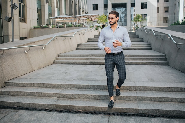 La imagen del hombre joven atractivo camina en pasos y mira a la izquierda. Él sostiene la taza de café en la mano izquierda. Guy usa gafas de sol. Se ve elegante.