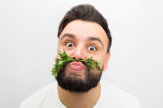 Imagen del hombre joven divertido que se coloca y que mira. Él hace muecas. Esta vez el chico pone el perejil entre los labios y la nariz.