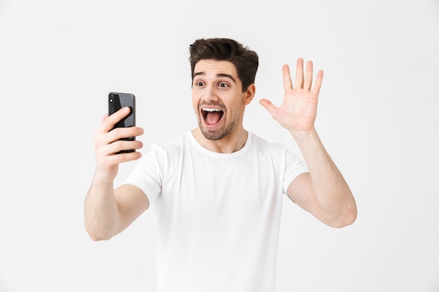 Imagen de hombre joven feliz emocionado posando aislada sobre pared blanca con teléfono móvil tomar un selfie saludando.