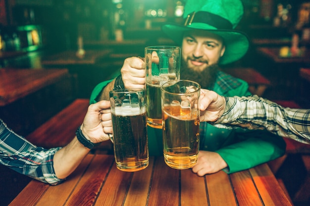 La imagen del hombre joven en el traje verde de St Patrick se sienta a la mesa con los amigos en pub. Sostienen jarras de cerveza juntas.