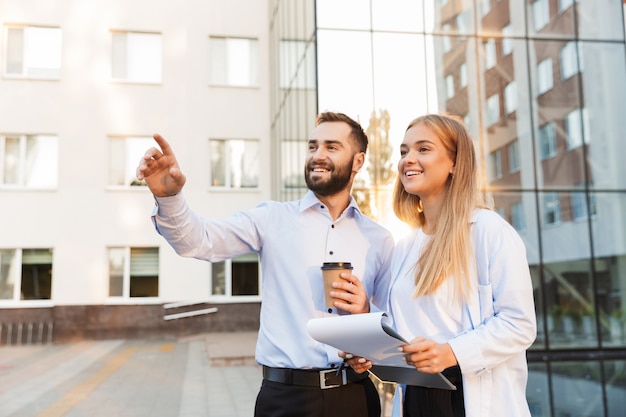 Imagen de un hombre y una mujer felices jóvenes empresarios afuera en la calle cerca del centro de negocios sosteniendo el portapapeles con planes de documentos y una taza de café apuntando mirando a un lado.