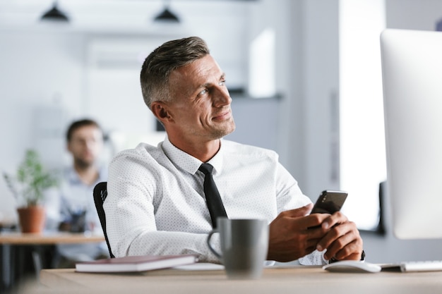 Imagen de hombre de negocios de 30 años con camisa blanca y corbata sentada en el escritorio en la oficina por computadora y sosteniendo el teléfono inteligente