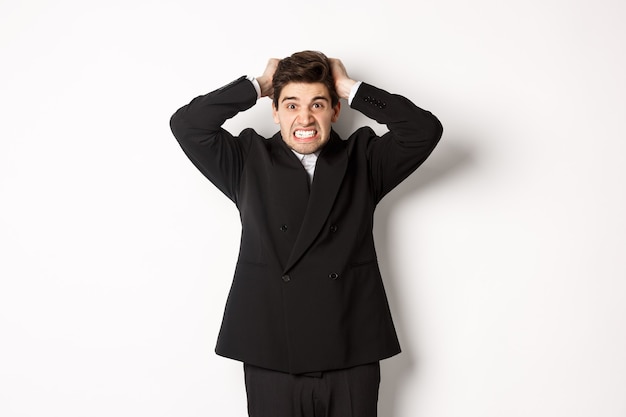 Imagen del hombre de negocios frustrado y enojado en traje negro, rasgando el pelo en la cabeza y haciendo muecas de enojo, de pie tenso contra el fondo blanco.