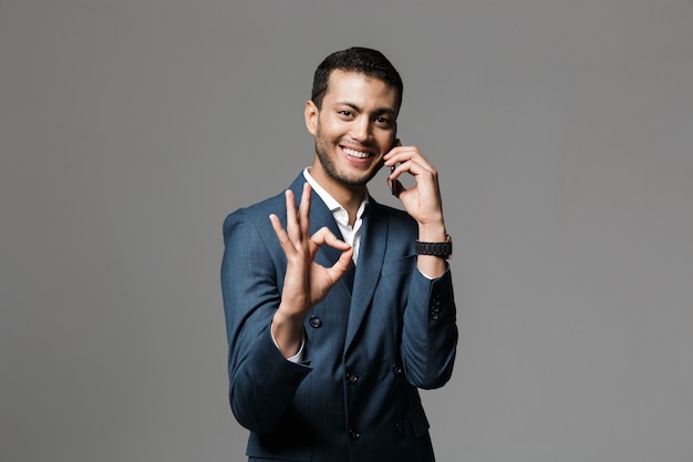 Imagen de un hombre de negocios joven sonriente feliz hermoso aislado sobre la pared gris de la pared que habla por teléfono móvil que muestra el gesto correcto.