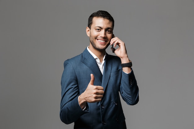 Imagen de un hombre de negocios joven sonriente feliz hermoso aislado sobre la pared gris de la pared que habla por teléfono móvil que muestra los pulgares arriba gesto.