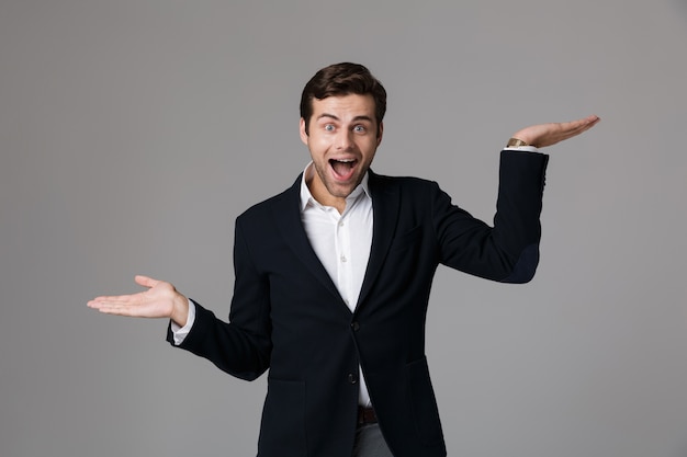 Imagen del hombre de negocios sorprendido de 30 años en traje formal gritando y levantando los brazos, aislado sobre la pared gris