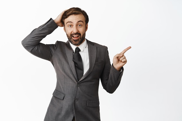 Imagen de un hombre de negocios sorprendido señalando con el dedo a la derecha mirando con incredulidad de pie con traje gris sobre fondo blanco