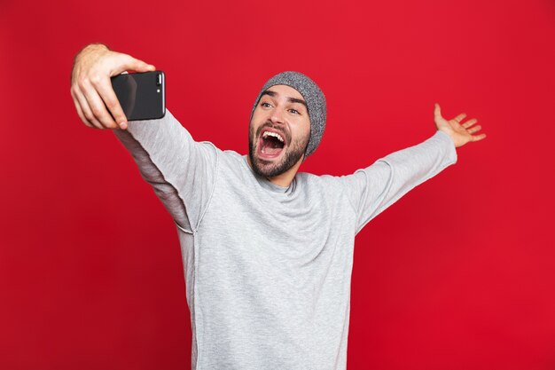 Imagen de hombre positivo de 30 años en ropa casual riendo mientras toma una foto selfie en un teléfono celular aislado