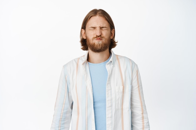 Imagen de un hombre rubio con barba que cierra los ojos, conteniendo la respiración, de pie con un pantalones blanco casual sobre fondo blanco. copia espacio