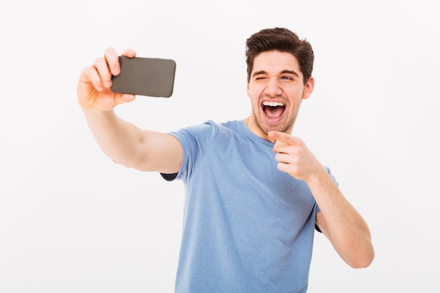 Imagen de hombre satisfecho con cabello castaño y cerdas sonriendo y señalando con el dedo a la cámara mientras toma selfie en teléfono móvil negro, aislado sobre la pared blanca