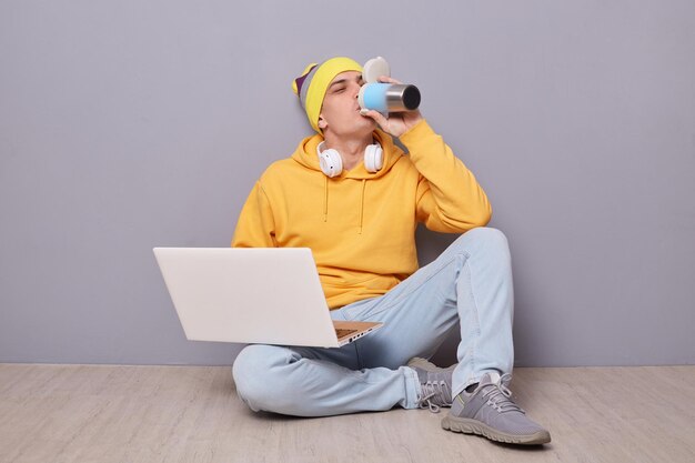 Imagen de un hombre sediento con gorro amarillo, sudadera con capucha y jeans posando contra una pared gris trabajando en una laptop sentada en el suelo y bebiendo café o té de un termo