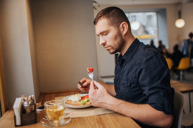 Una imagen del hombre sentado a la mesa y comiendo. Él tiene un trozo de tomate en el tenedor. Guy lo está mirando con mucha seriedad.