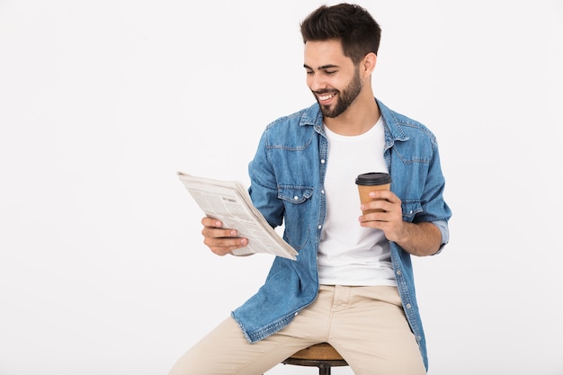 Imagen de hombre sonriente guapo bebiendo café para llevar y leyendo el periódico mientras está sentado en una silla aislada sobre pared blanca