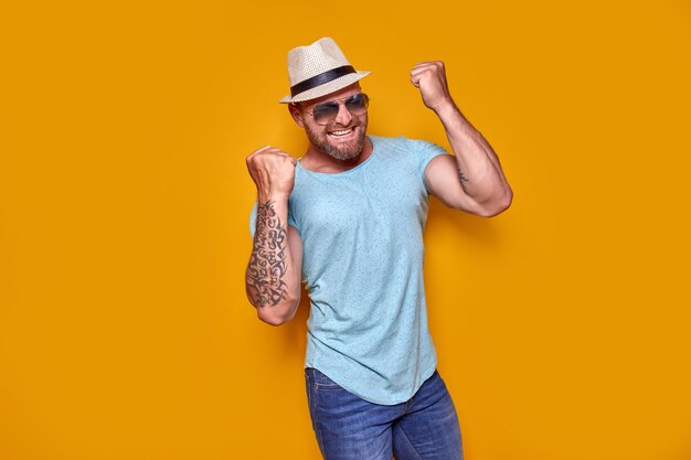 Foto imagen de hombre tatuado con barba emocionado con gafas de sol y sombrero haciendo gesto de ganador aislado ba amarillo ...