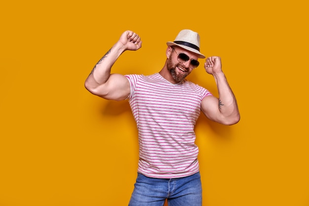 Imagen de hombre tatuado con barba emocionado con gafas de sol y sombrero haciendo gesto de ganador aislado ba amarillo ...