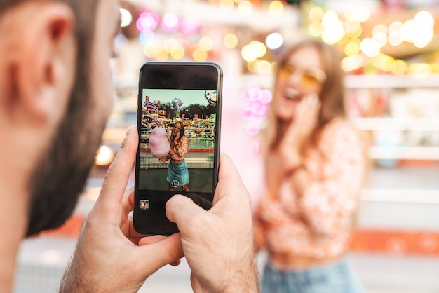 Imagen de un hombre toma una foto de su mujer feliz caminando al aire libre en el parque de atracciones por teléfono móvil.