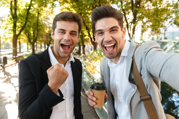 Foto imagen de hombres de negocios alegres en trajes tomando foto selfie en teléfono celular, mientras camina al aire libre a través del parque verde con café para llevar