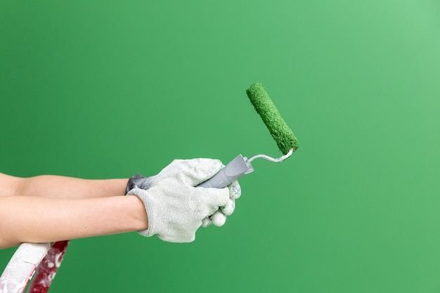 Imagen horizontal cercana de manos con guantes de trabajo sosteniendo un rodillo de tamaño medio con pintura verde frente a la pared verde