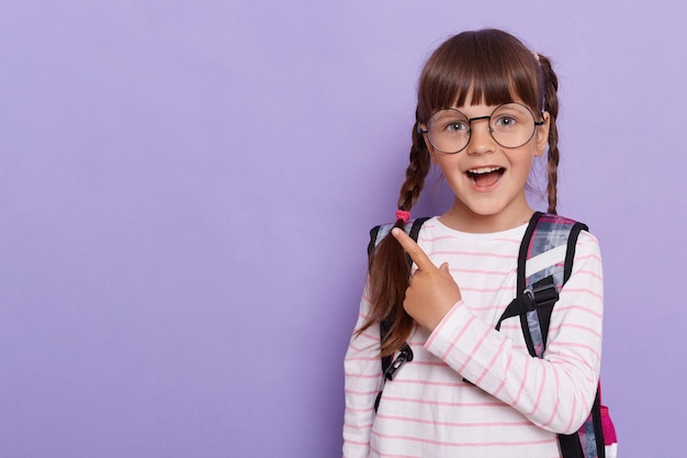 Imagen horizontal de una colegiala emocionada señalando con los dedos a un lado mostrando espacio de copia para texto promocional con pantalones a rayas y gafas aisladas en un fondo violeta