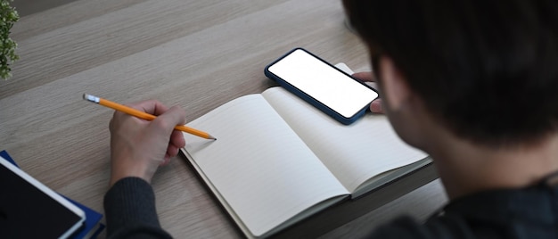 Imagen horizontal del hombre tomando nota en el cuaderno vacío y usando el teléfono móvil en la mesa de madera.