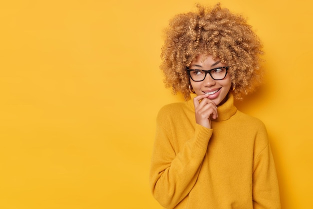 Imagen horizontal de una joven encantadora y soñadora que mantiene el dedo en los labios mira hacia otro lado con una curiosa expresión feliz vestida con un cómodo puente y espectáculos aislados sobre un espacio de copia de fondo amarillo