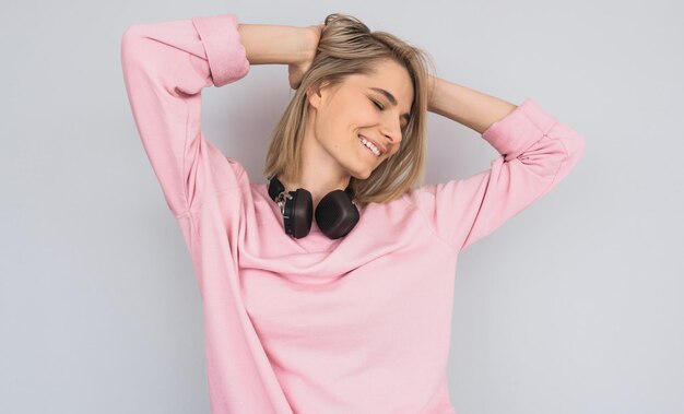 Imagen horizontal de una joven feliz y sonriente con suéter rosa bailando con auriculares en el cuello escuchando música favorita aislada en el fondo del estudio blanco Copiar espacio para texto publicitario