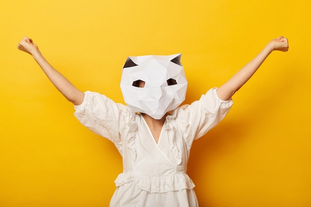 Imagen horizontal de una mujer alegre y optimista con vestido y máscara de gato de papel posando aislada sobre un fondo amarillo de pie con los brazos levantados bailando mostrando triunfo hurra