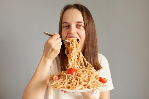 Imagen horizontal de una mujer delgada y hermosa con el pelo oscuro comiendo pasta mirando a la cámara disfrutando de comida poco saludable cenando posando aislada sobre un fondo gris