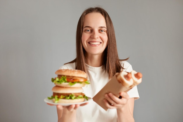 Imagen horizontal de una mujer sonriente y satisfecha con el pelo castaño aislada en un fondo gris sosteniendo perritos calientes y hamburguesas americanas disfrutando de comida rápida