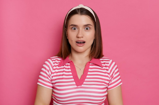 Imagen horizontal de una mujer sorprendida con una camiseta a rayas y una banda para el cabello posando aislada sobre un fondo rosado mirando la cámara con ojos grandes y boca abierta