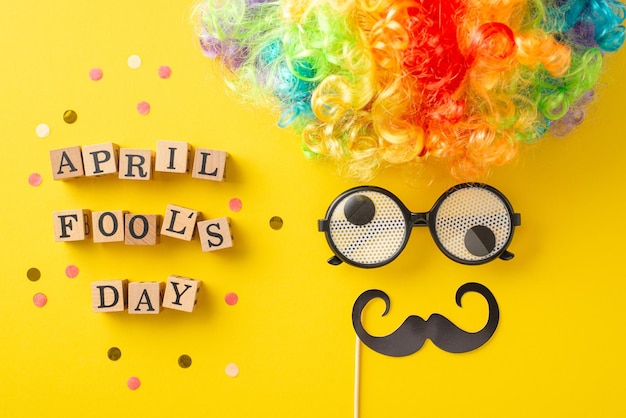Imagen instantánea de letras de madera que escriben confeti del día de los tontos de abril con accesorios de fiesta como payasos, pelucas, gafas y bigote que crean una cara humorística en una superficie amarilla brillante
