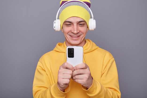 Imagen interior de un amable hombre caucásico con sombrero de beanie y capucha informal amarilla sosteniendo un teléfono inteligente en las manos revisando las redes sociales escribiendo mensajes aislados sobre un fondo gris