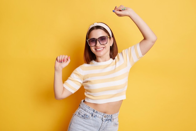 Imagen interior de una atractiva mujer extremadamente feliz con pantalones a rayas y jeans levantando los brazos y bailando con buenas noticias y un gran espíritu posando aislada sobre un fondo amarillo