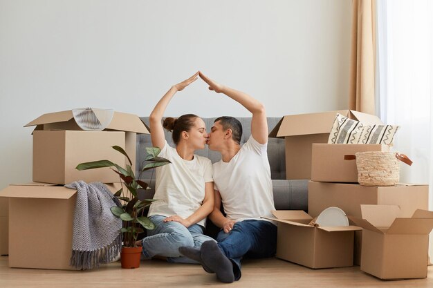 Imagen interior de una feliz y encantadora pareja romántica sentada en el suelo rodeada de cajas de cartón durante la reubicación en un nuevo apartamento con camisetas blancas haciendo techo sobre sus cabezas con las manos