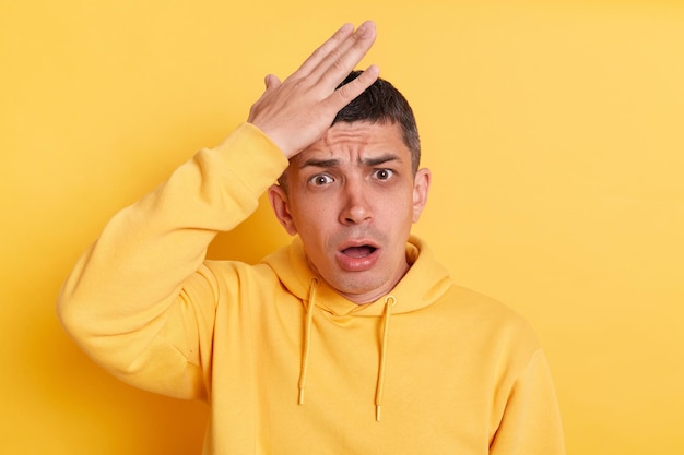 Imagen interior de un hombre asustado conmocionado con una capucha de estilo informal posando aislado sobre fondo amarillo mirando la cámara con la boca abierta manteniendo la mano en la frente